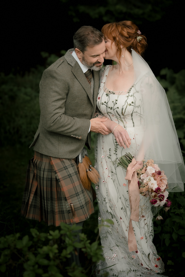 Bride and groom embracing outdoors, holding bouquet during wedding portraits.