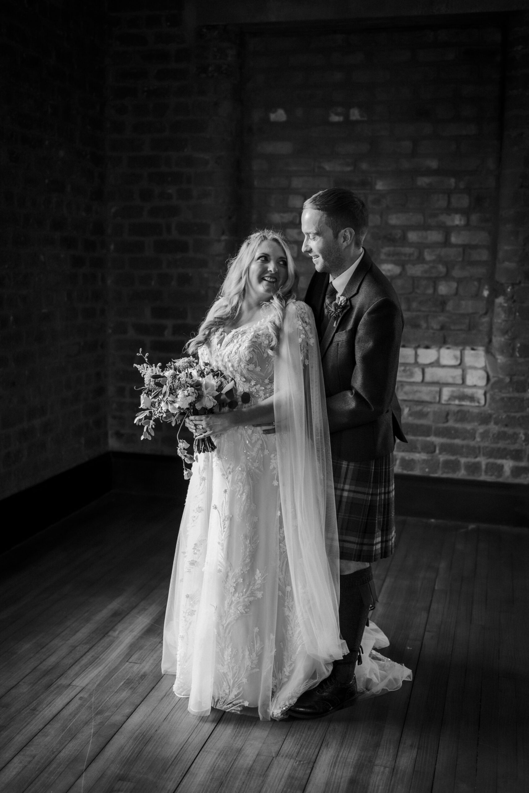 Bride holding bouquet beside groom in a brick-walled room during a wedding portrait.