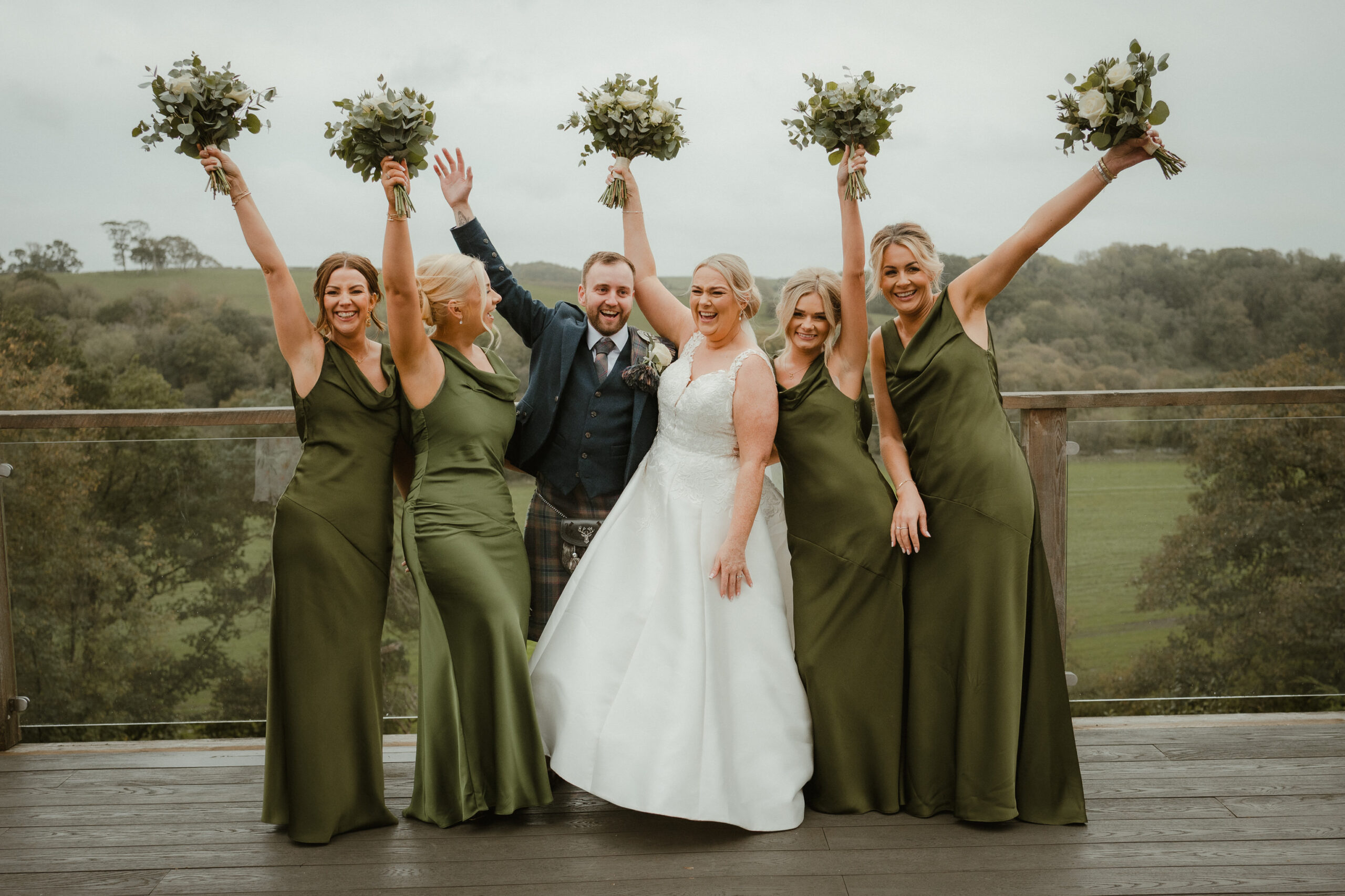 Bride, groom and bridesmaids in olive dresses raising their bouquets on the deck at Enterkine House, Ayrshire