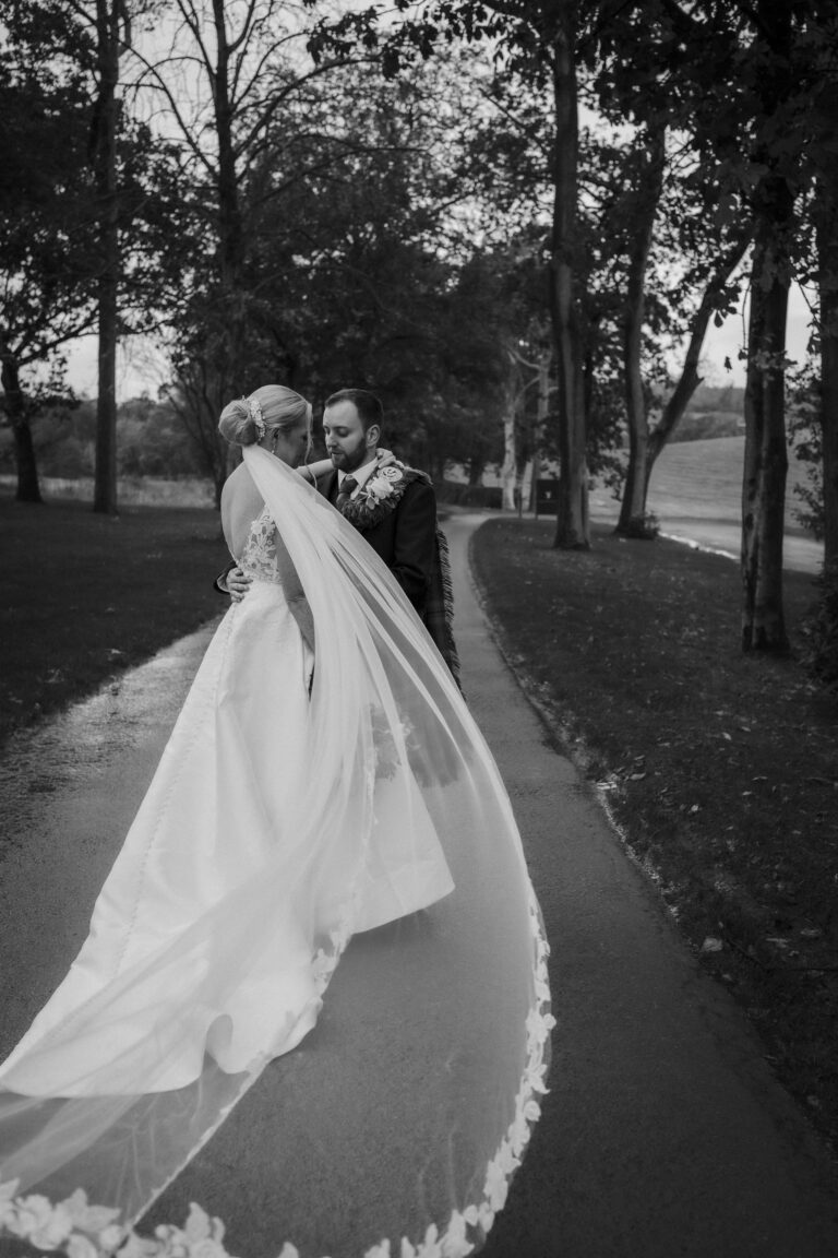 Bride and groom on the tree-lined drive at Enterkine House, her long veil trailing behind
